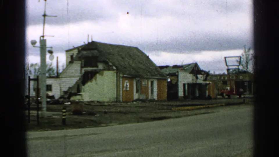 1965 Minnesota frame showing the damaged house from a slightly shifted roadside angle with muted 8mm color.