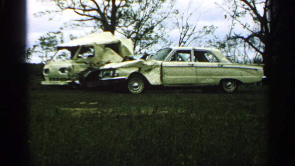 1965 Minnesota frame showing a damaged vintage car and camper in a grassy area with bare trees behind them.