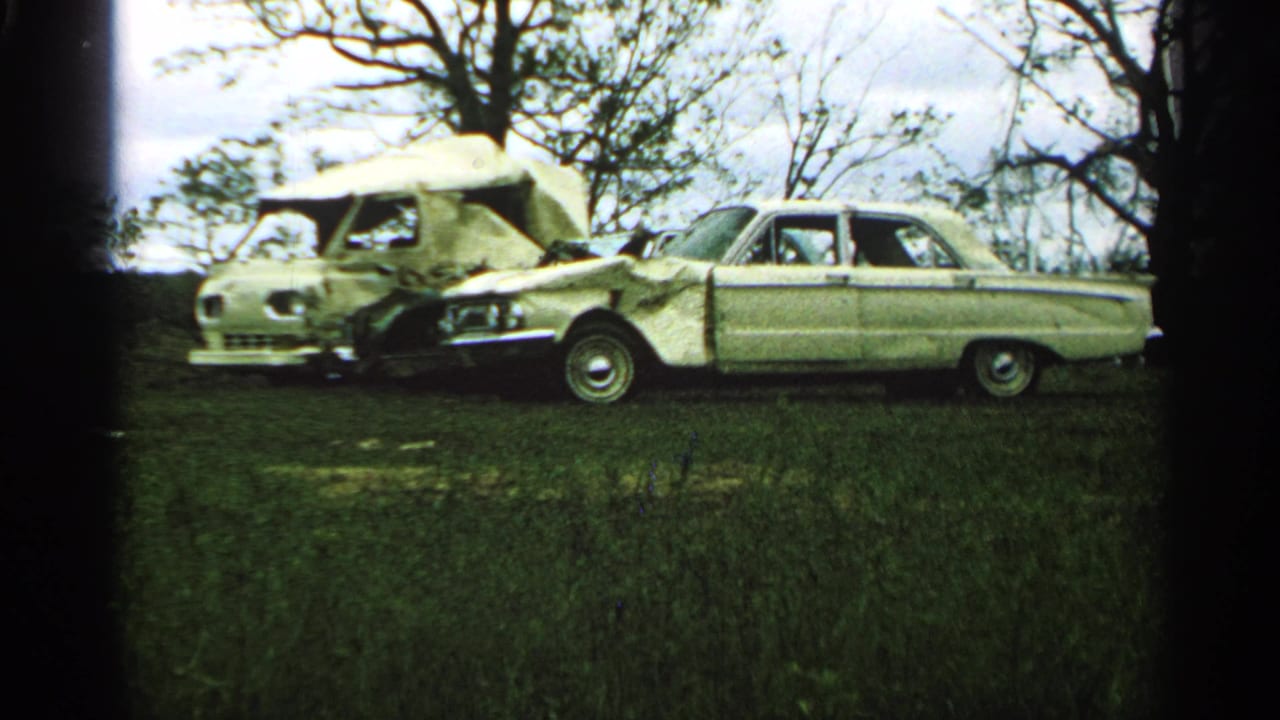1965 Minnesota home-movie frame with a crushed-front sedan beside a damaged camper van in an open grassy area.