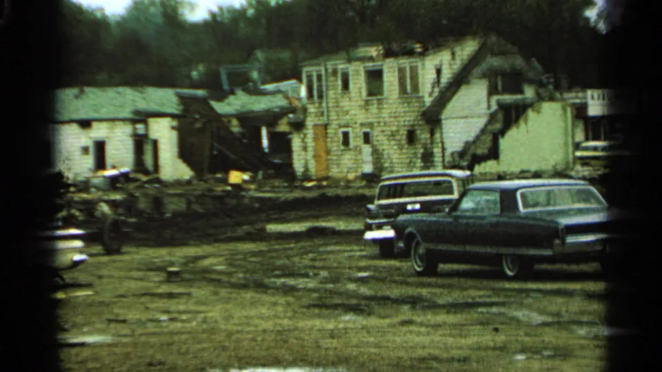 1965 Minnesota 8mm frame showing a damaged house, debris-strewn yard, and parked 1960s cars in the foreground.