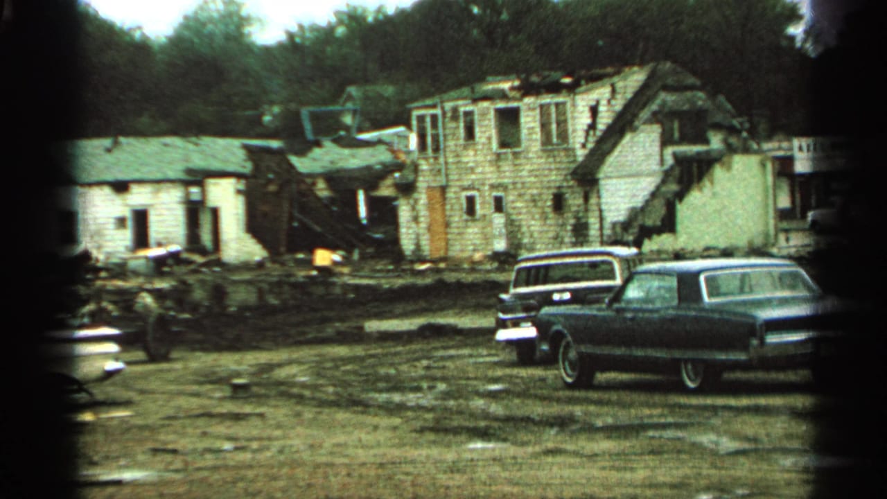 1965 Minnesota 8mm frame showing damaged rural buildings, muddy debris, and parked mid-century cars in overcast light.
