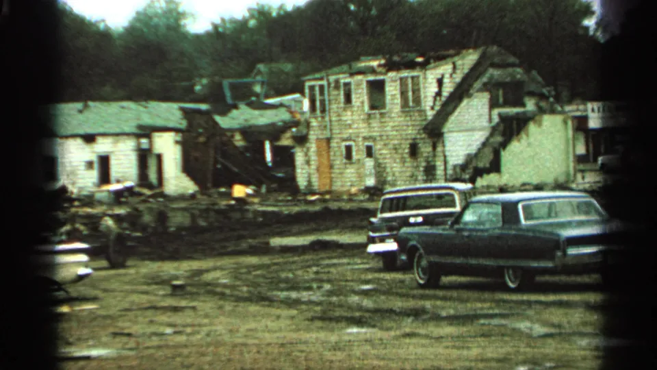 1965 Minnesota 8mm frame showing damaged rural buildings, muddy debris, and parked mid-century cars in overcast light.