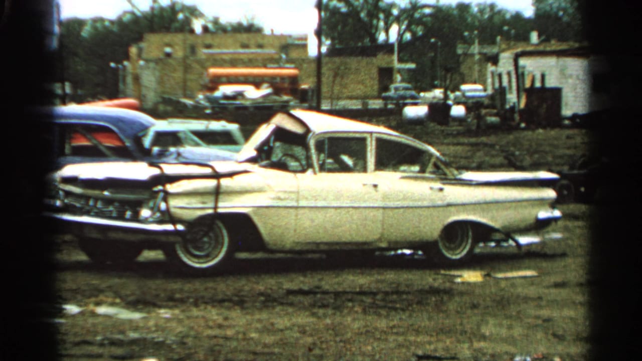 1965 Minnesota 8mm frame with a white mid-century sedan, crumpled hood, muddy ground, brick buildings, and parked cars.