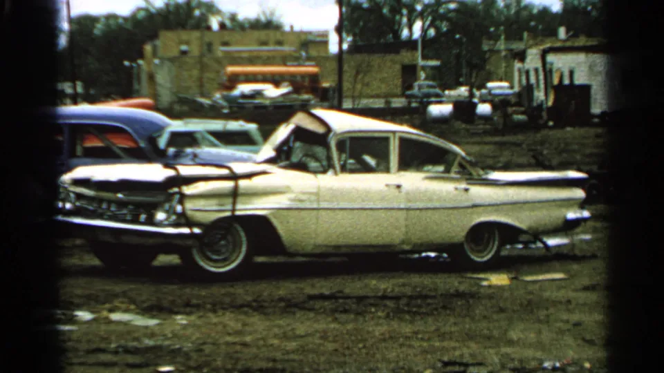 1965 Minnesota 8mm frame centered on a white damaged sedan with chrome trim, whitewall tires, muddy field, and background buildings.
