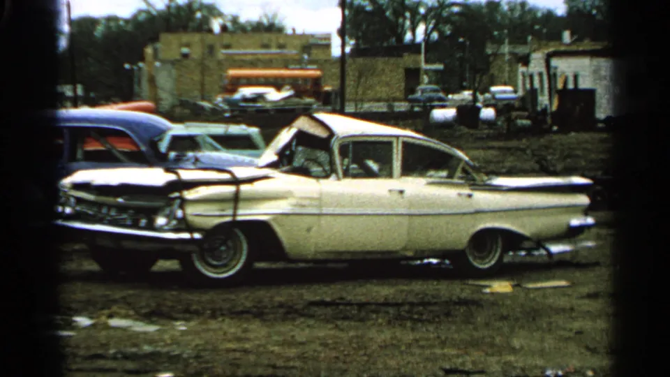 1965 Minnesota 8mm frame showing a damaged white sedan in profile, with muddy debris, brick buildings, and other parked vehicles behind.