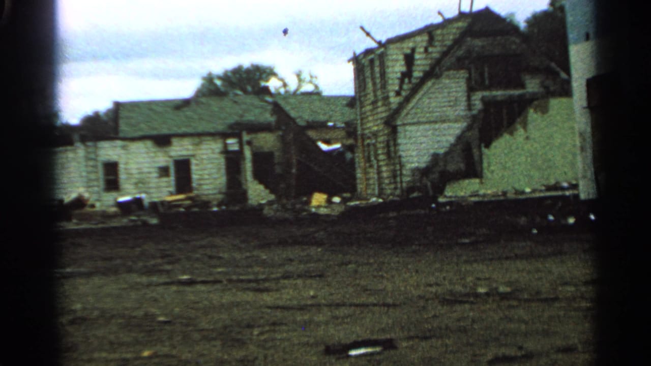 Minnesota 1965 home-movie frame showing a severely damaged wooden house with siding, framing, and debris scattered outside.