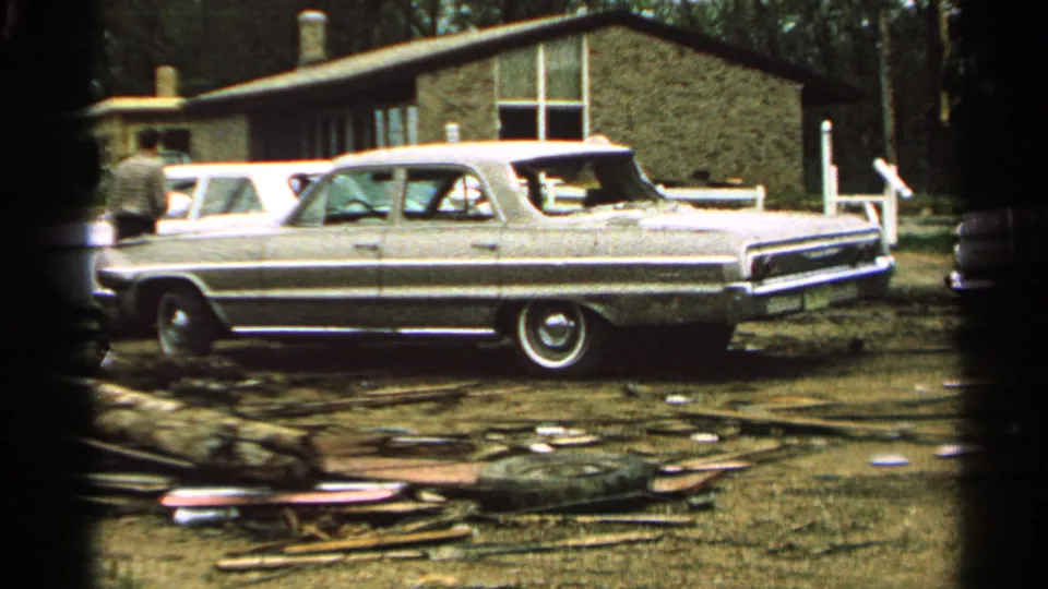 Minnesota 1965 home-movie frame showing a long American sedan parked by debris, with an adult standing near a low brick building.