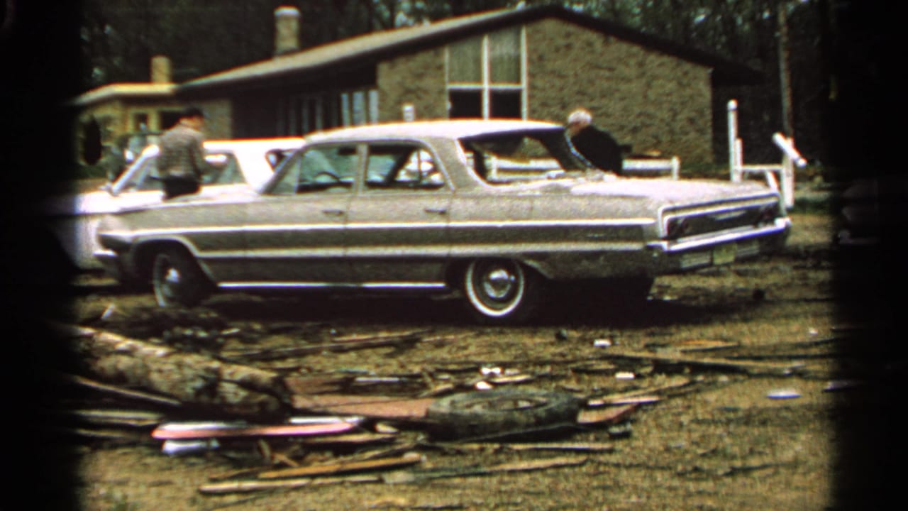 1965 Minnesota home-movie frame showing two adult men near a large sedan beside a damaged-looking house and scattered yard debris.