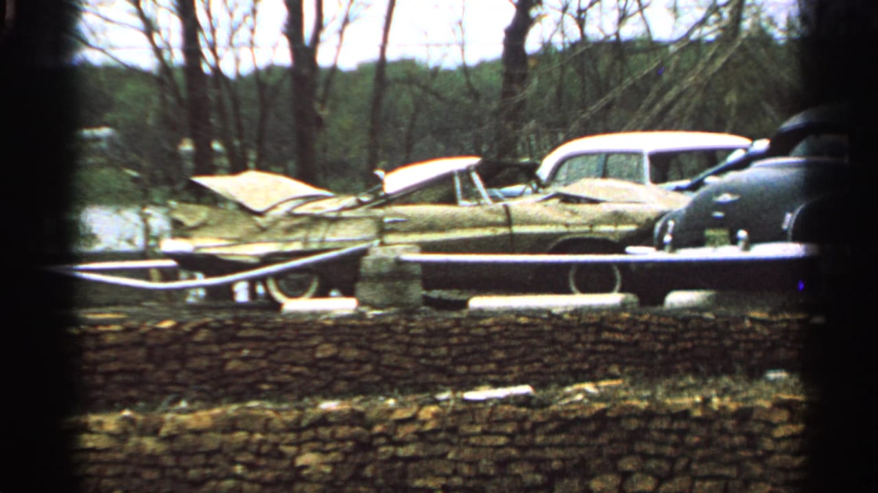 1965 Minnesota 8mm frame showing abandoned cars behind a stone wall, including a light-colored sedan with its hood raised.