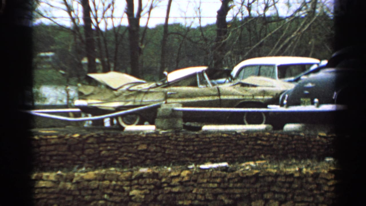 1965 Minnesota home-movie view of weathered cars near a stone wall, partly obscured by dark foreground framing.