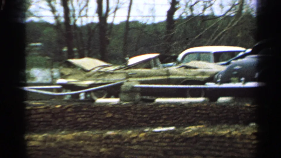 1965 Minnesota 8mm frame with a blurred view of abandoned cars, a low stone wall, and leafless trees.