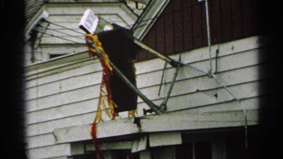 1965 Minnesota 8mm frame showing a damaged house exterior with broken antenna rods, siding, and torn hanging fabric.