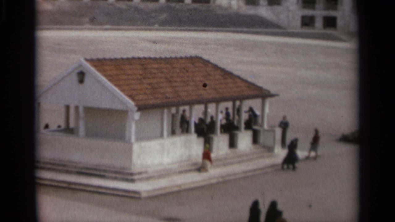 White pavilion with a terracotta roof and visitors standing on steps in an open plaza.