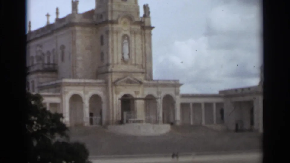 1965 8mm view of the Nazaré basilica exterior, framed with trees at left and a long colonnade to the right.