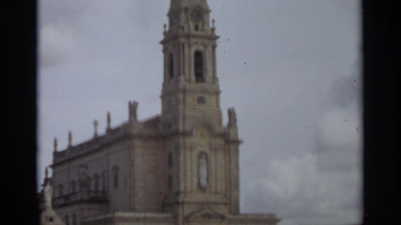 1965 8mm frame from Nazaré, Portugal, showing an ornate church facade and tall bell tower in soft focus beneath an overcast sky.