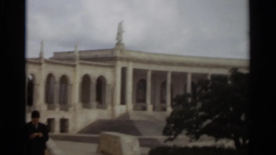 1965 8mm frame in Nazaré showing a columned church structure, a large foreground tree, and a small visitor near the lower left edge.