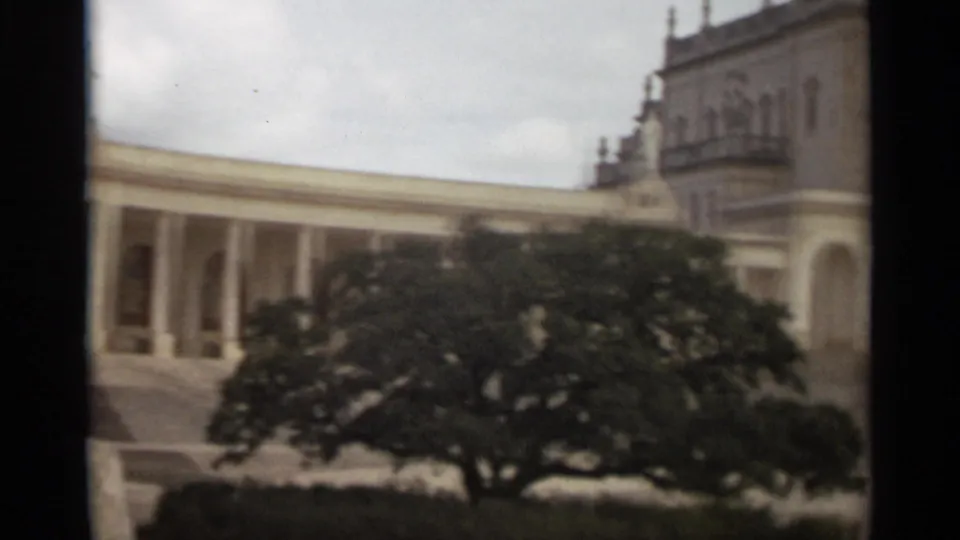 1965 Nazaré home-movie frame with a church colonnade, large leafy tree, and partial bell tower at the right under cloudy sky.