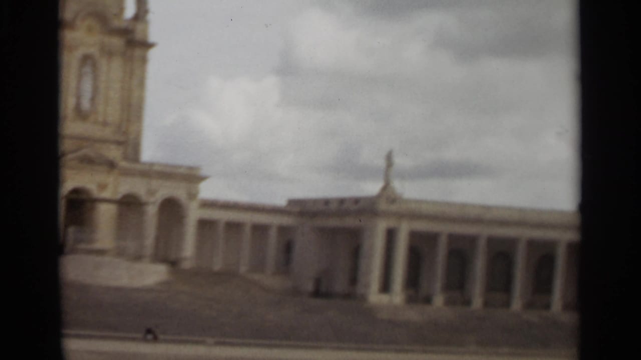 1965 Nazare home-movie frame showing a bell tower, columns, steps, and pale facade beneath a cloudy sky.