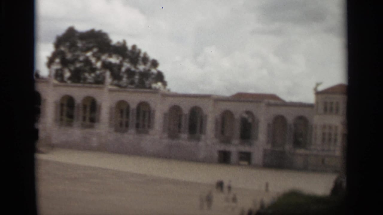 1965 Nazaré home-movie frame showing a pale arched architectural facade, red roof sections, open courtyard, and small distant pedestrians.