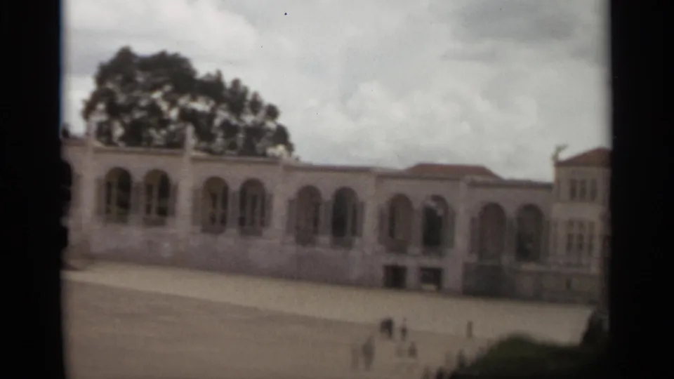 1965 Nazaré home-movie frame showing a pale arched architectural facade, red roof sections, open courtyard, and small distant pedestrians.