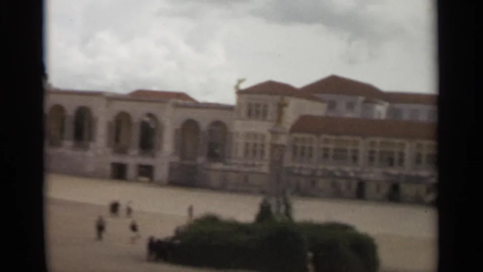 The camera pans across the Nazaré complex, showing arched windows, red-tiled roofs, a broad paved area, shrubs, and small figures walking.