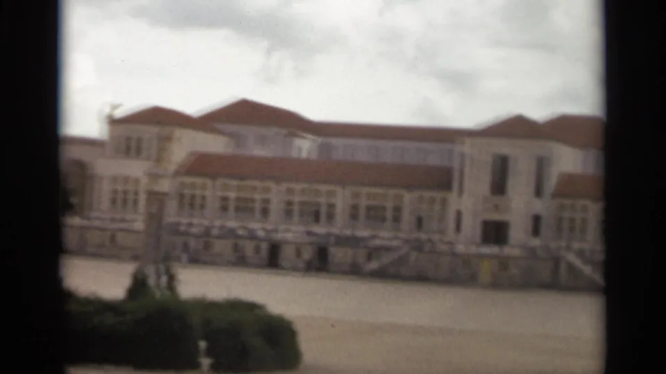 Mid-pan frame of the Nazaré architectural complex with a long windowed wing, red roofline, pale walls, courtyard paving, and foreground greenery.
