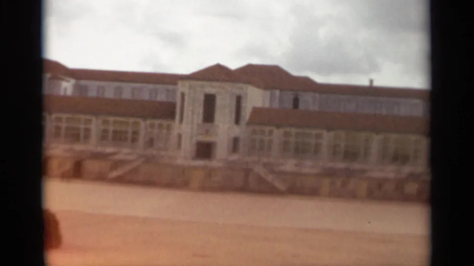 Final Nazaré frame showing the red-roofed complex, broad courtyard, stair details, cloudy sky, and a warm exposure flare at the lower left.