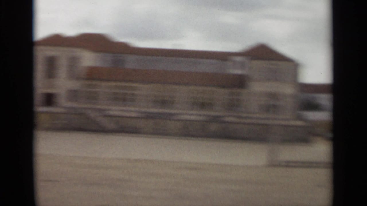 Blurred exterior of a large pale building with a reddish roof across an open area.