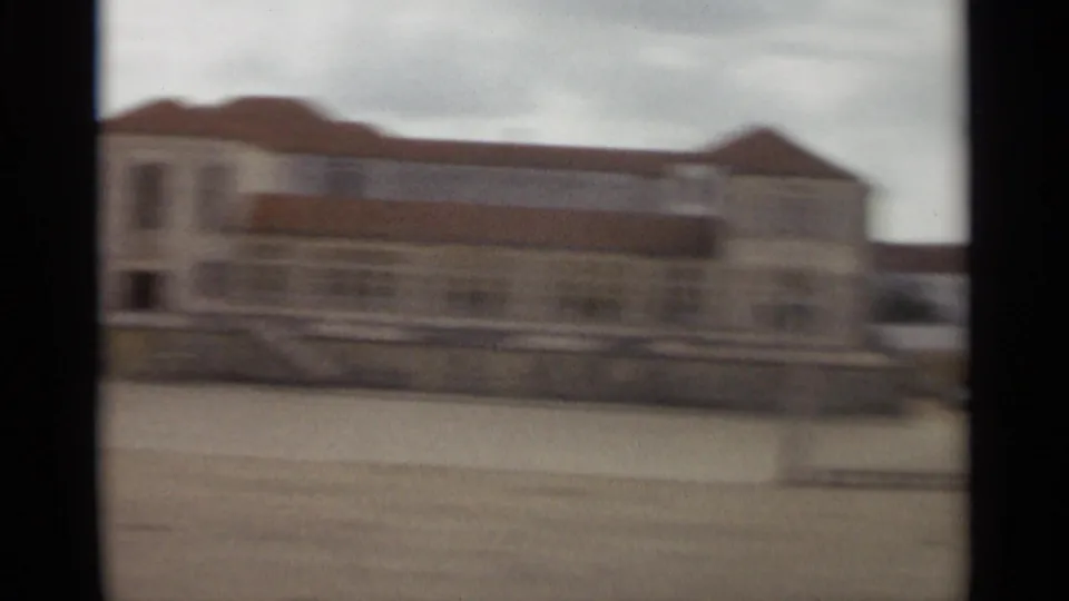 Soft 8mm view from Nazare, Portugal, showing a pale building with a reddish roof across an open foreground in 1965 footage.