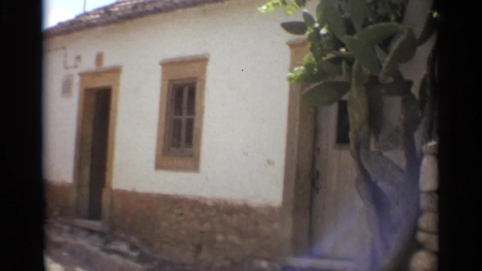 A whitewashed building in Nazare with doorway, narrow window, cactus growth, and stonework appears in soft 1965 8mm footage.