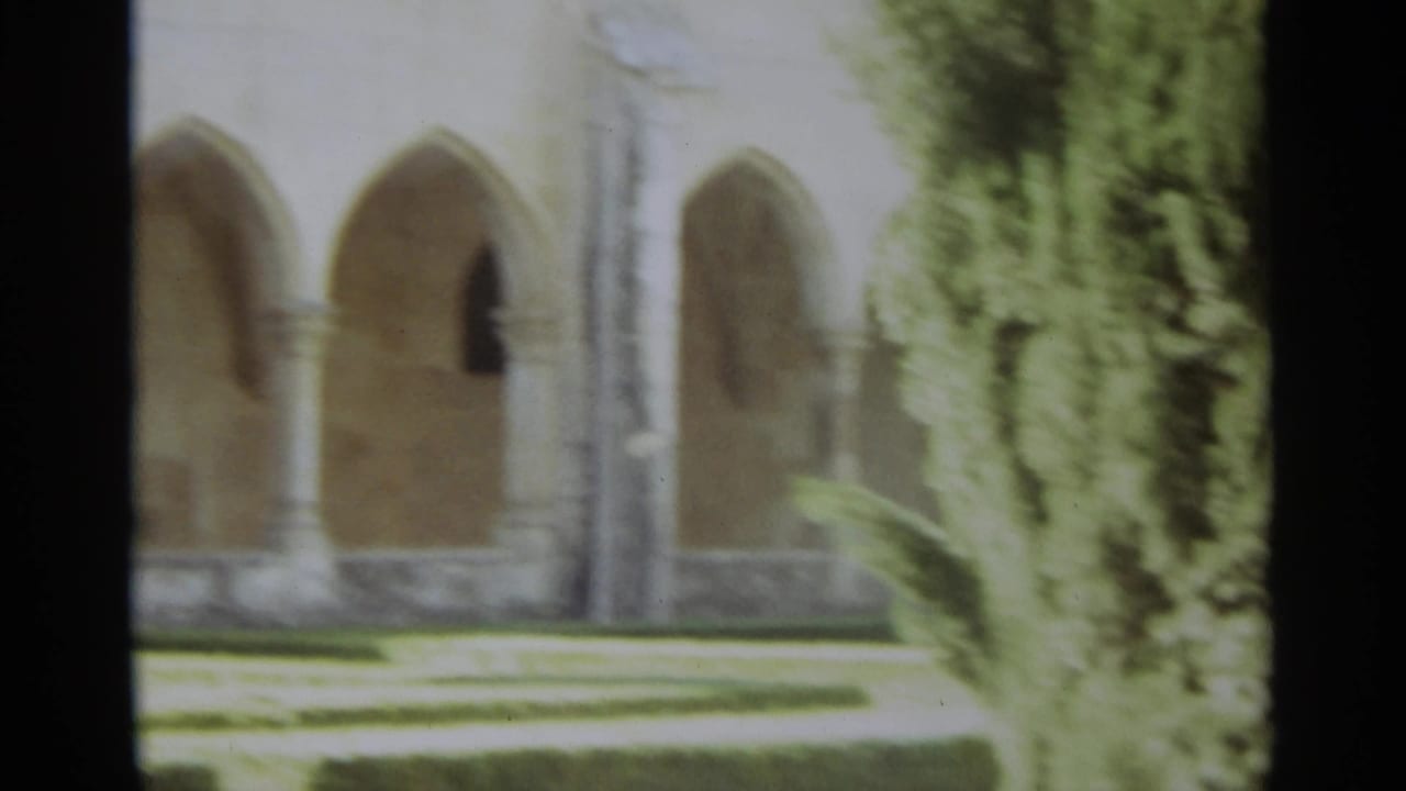 1965 Nazaré, Portugal 8mm frame showing a religious courtyard arcade behind trees and low hedges.