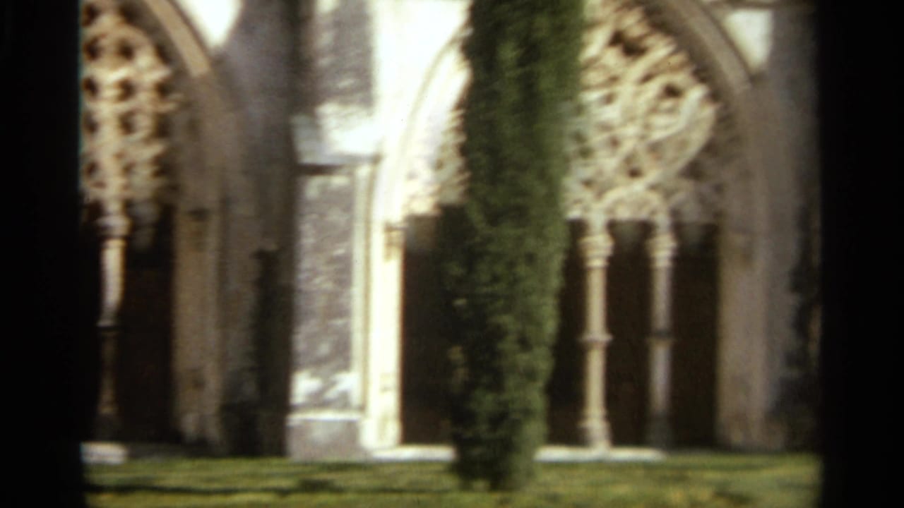 1965 Nazare, Portugal 8mm frame showing a church cloister with pointed arches, ornate tracery, stone columns, lawn, and a tall hedge.