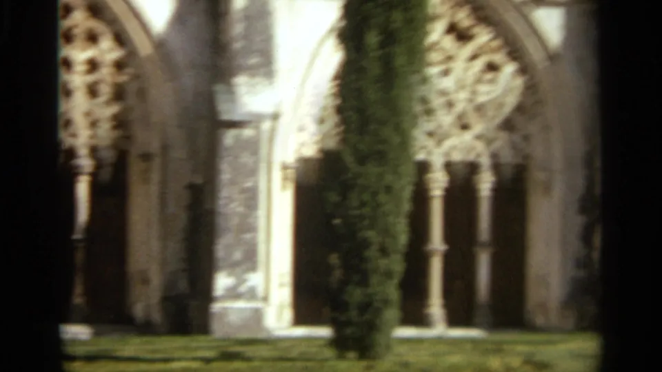 1965 Nazare, Portugal 8mm frame showing a church cloister with pointed arches, ornate tracery, stone columns, lawn, and a tall hedge.