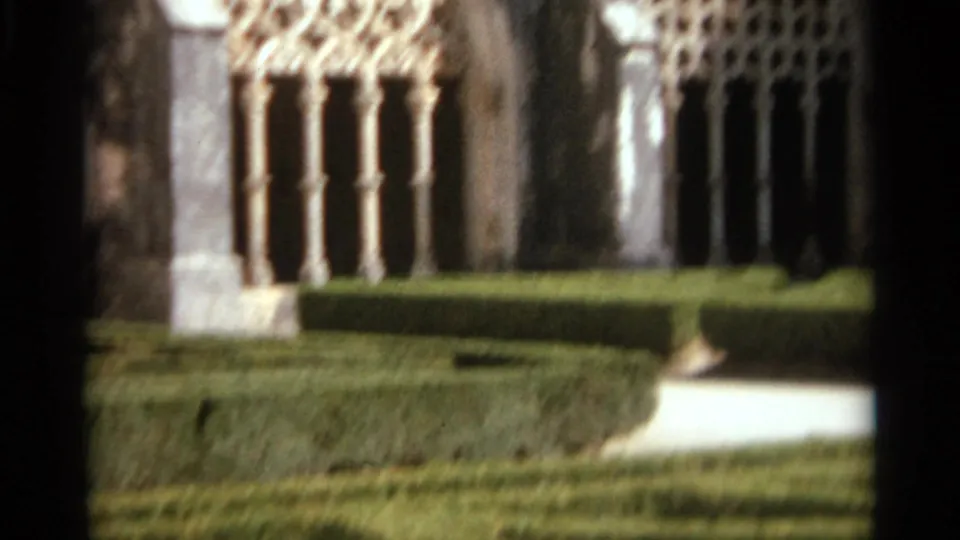 1965 Nazare, Portugal 8mm frame showing a cloister corner with carved arches, columns, manicured hedges, lawn, and a pale path.