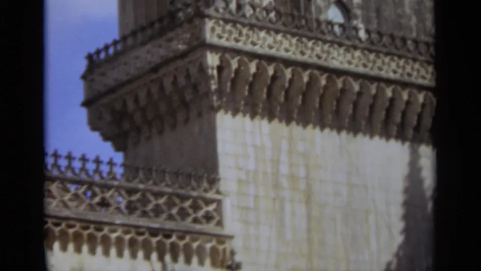 1965 Nazaré, Portugal 8mm frame of an ornate church tower corner with carved stone trim, parapet rail, and clear blue sky.