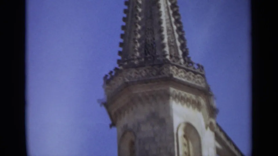 1965 Nazaré, Portugal 8mm frame capturing a tall church spire, decorative upper tower, pale stone facade, and clear sky.