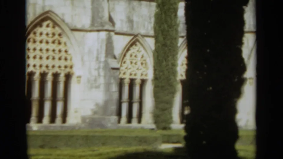 Wider 1965 home-movie frame showing two Gothic-style church windows, trimmed grass and a dark vertical tree or shadow near the facade.