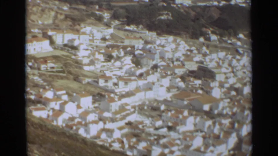 Elevated 1965 Nazaré, Portugal town view with white buildings, rooftops, roads, and a hillside beyond.