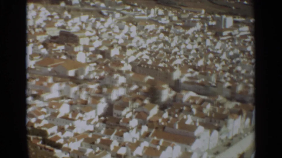 1965 Nazare aerial footage frame showing compact rooftops, pale facades, narrow streets, and a curved edge road.
