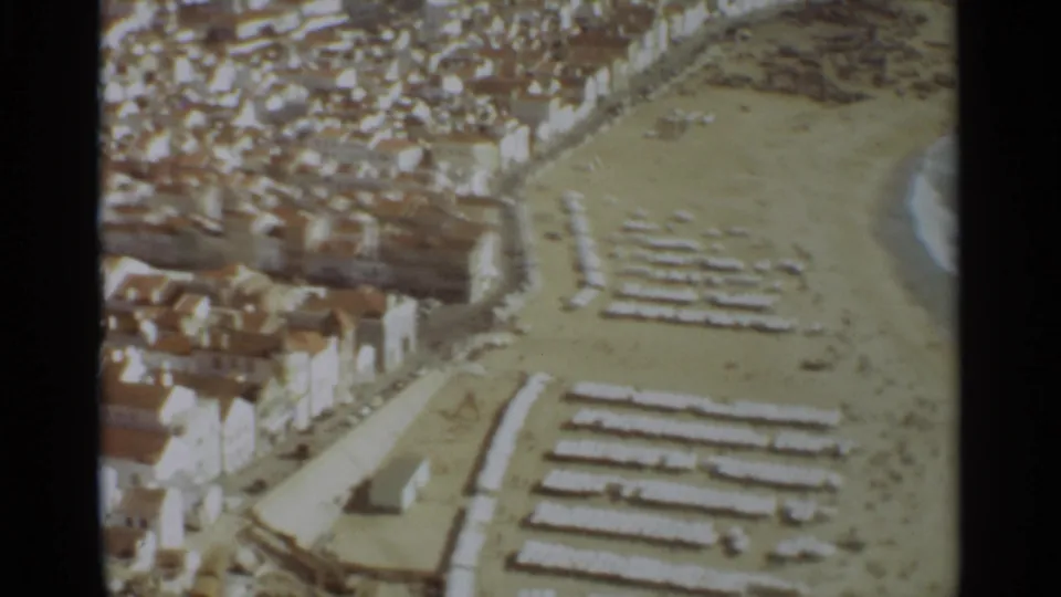 1965 aerial 8mm frame of Nazare beach with arranged umbrellas or loungers, town buildings, and surf at the edge.
