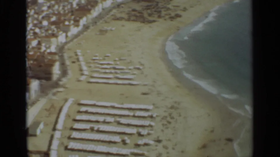 Nazare coastline in 1965, viewed from above with rows of beach umbrellas or loungers on sand beside blue-green surf.