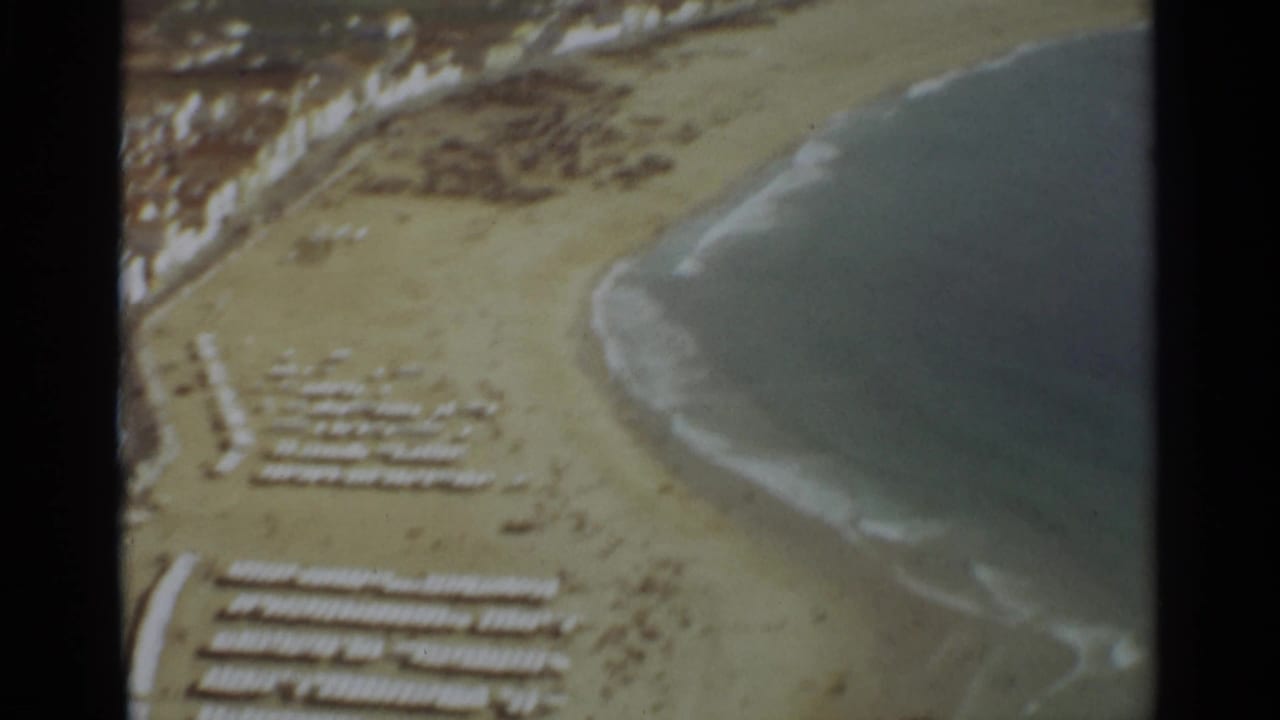 1965 8mm aerial view over Nazare, Portugal, showing a curved sandy beach, organized white beach furniture, pale waterfront buildings, and waves reaching the shore.