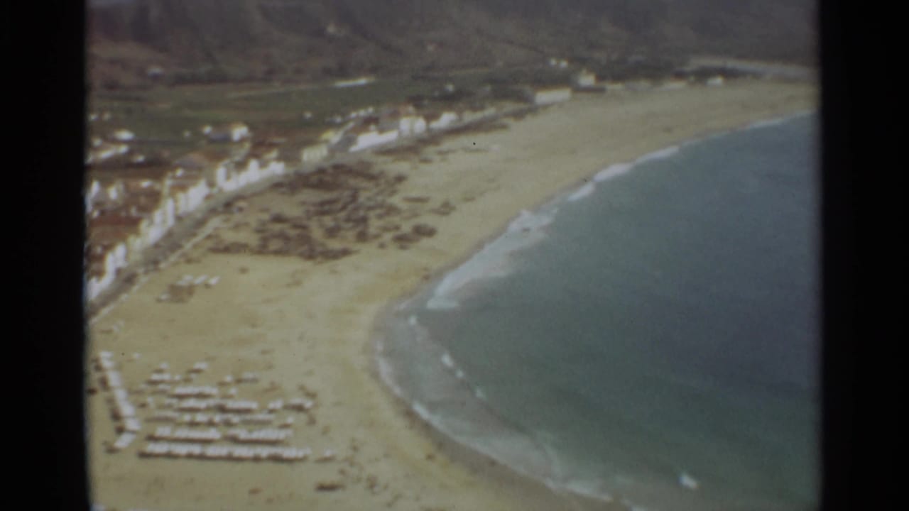 Aerial coastline view with a sandy beach, surf, waterfront buildings, and hills inland.