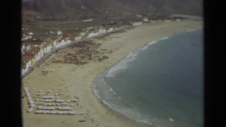 1965 8mm aerial frame of the Nazare shoreline, with the Atlantic surf curving along the sand, beach furniture below, white buildings near the water, and low hills behind.
