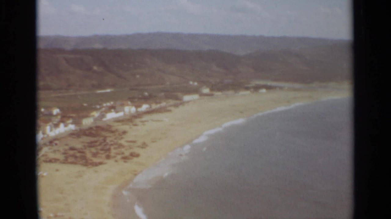 Aerial view across a beach toward waterfront buildings and hills, with surf at the lower right.