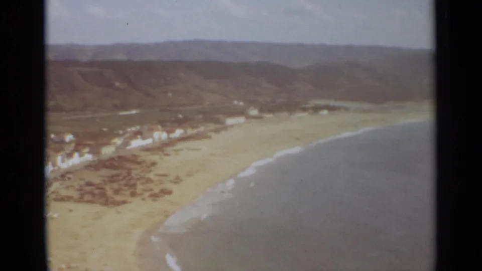 1965 8mm aerial view from Nazare, Portugal, looking across the beach toward pale town buildings and inland hills while the surf line curves through the foreground.