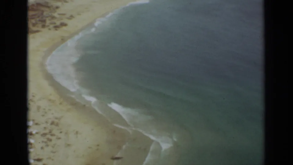 1965 8mm aerial frame over the Nazare coast, emphasizing blue-green Atlantic water, white wave lines, and the edge of the sandy beach below.