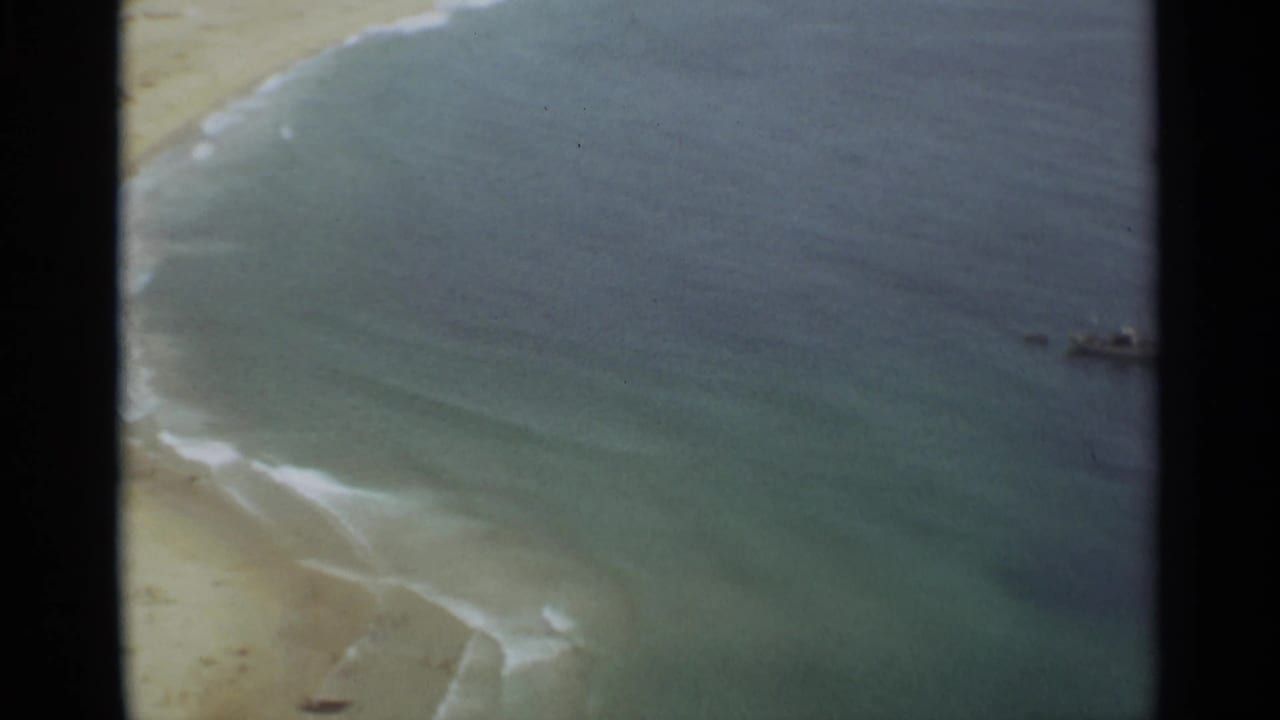1965 Nazare, Portugal home-movie frame showing sandy shoreline, gentle surf, and blue-green coastal water.