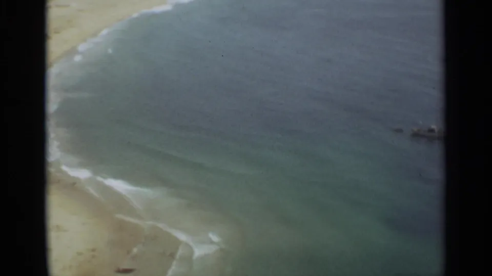 1965 Nazare, Portugal home-movie frame showing sandy shoreline, gentle surf, and blue-green coastal water.