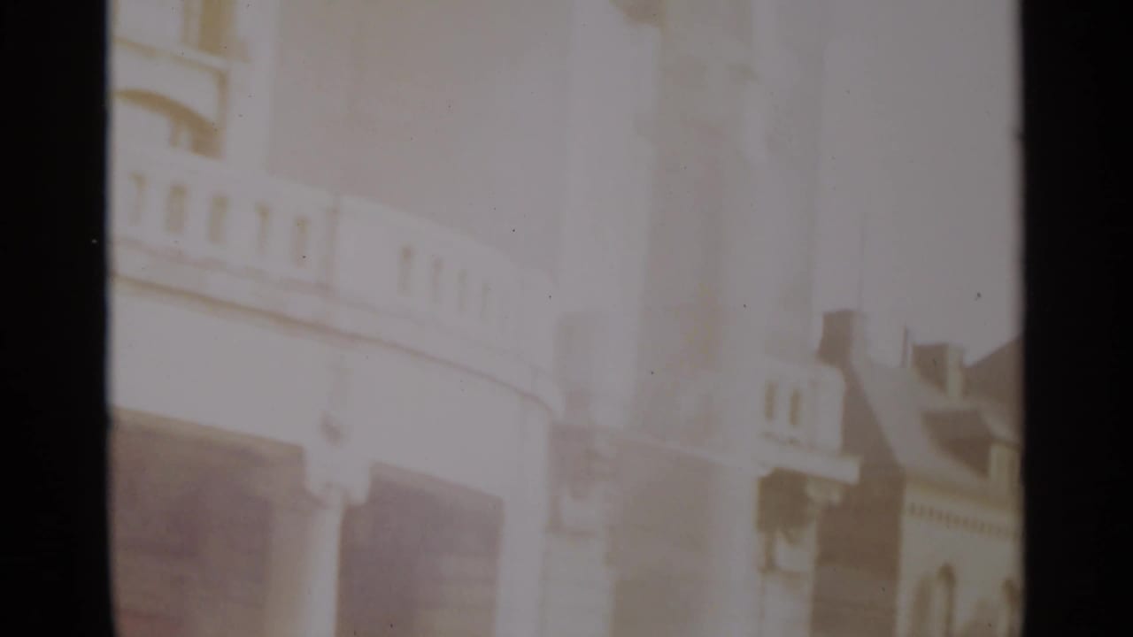 1965 Nazare frame with a washed, bright building front, balcony railings, dark openings, and a neighboring roofline at the right edge.